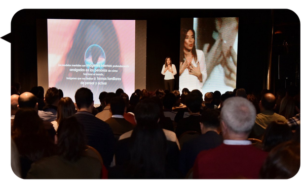 Natalia De Vita impartiendo una conferencia ante un gran público, con una presentación visual proyectada en la pantalla.