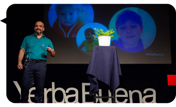 Hugo Quintana en el escenario de TEDx Yerba Buena, con imágenes de niños proyectadas en la pantalla y una planta sobre una mesa cubierta con tela azul.