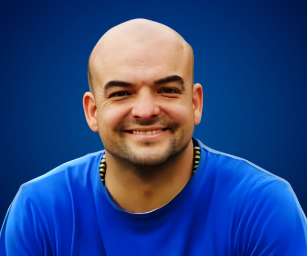 Retrato de Hugo Quintana sonriendo, vistiendo una camiseta azul con un fondo azul.