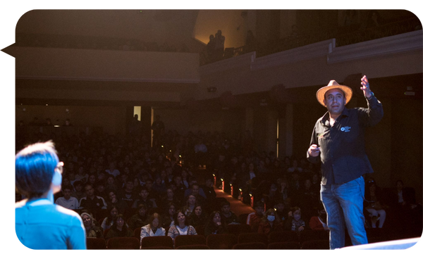 Gaspar Contreras en el escenario de un teatro, vistiendo camisa negra y sombrero, interactuando con la audiencia mientras expone sobre bienestar y felicidad organizacional.