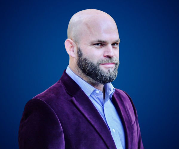 Fotografía de Alejandro Lanuque, retrato profesional de medio cuerpo con fondo azul, vistiendo saco morado de terciopelo y camisa celeste, con expresión confiada.